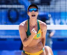 #ParaQuemNaoPodeVer A imagem mostra uma jogadora de vôlei de praia em ação. Ela está vestindo um biquíni amarelo com detalhes verdes e o símbolo da bandeira do Brasil. A jogadora está usando óculos escuros e um boné preto. Ela parece estar comemorando ou incentivando, com a boca aberta e uma expressão de entusiasmo. Ao fundo, há uma rede de vôlei e um cenário azul, típico de uma quadra de vôlei de praia.
