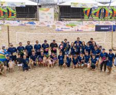 #ParaQuemNaoPodeVer A imagem mostra um grupo grande de pessoas posando para uma foto em uma quadra de areia, possivelmente de vôlei de praia. Todos estão vestindo camisetas azuis e alguns seguram bolas coloridas. Ao fundo, há uma estrutura com várias faixas e banners, que parecem ser de patrocinadores ou eventos, com logotipos e textos.