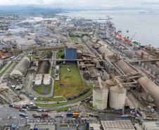 #ParaQuemNaoPodeVer A imagem mostra uma foto aérea de uma área portuária. Na imagem, é possível ver várias estruturas grandes, como tanques cilíndricos e prédios industriais, além de áreas verdes ao redor. Há também várias estradas e veículos visíveis.