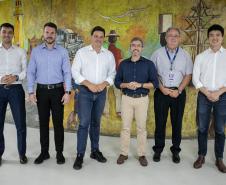#ParaQuemNaoPodeVer A imagem mostra uma foto de seis homens em pé, posando para a câmera. Eles estão em duas fileiras, três na frente e três atrás. Todos estão vestidos de forma formal, com camisas sociais e calças. O fundo da foto é uma pintura mural colorida que parece representar uma cena cultural ou histórica.
