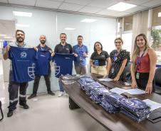 #ParaQuemNaoPodeVer A imagem mostra um grupo de pessoas em um ambiente de escritório. Na parte superior da imagem, há quatro homens em pé, segurando camisetas azuis com um logotipo branco. Eles estão sorrindo e parecem estar participando de algum tipo de evento ou celebração. Na parte inferior da imagem, há três mulheres e um homem em pé atrás de uma mesa, que está coberta com várias pilhas de camisetas azuis dobradas. As mulheres estão sorrindo e também parecem estar participando do mesmo evento.