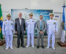 #ParaQuemNaoPodeVer A imagem mostra cinco homens posando para uma foto em frente a uma bandeira do Brasil e uma bandeira da Marinha do Brasil. Três deles estão vestidos com uniformes brancos de oficiais da Marinha, com chapéus brancos e insígnias no peito. Os outros dois homens estão usando ternos. Ao fundo, há um banner azul com o texto "13 de Dezembro" e "Dia do Marinheiro". A foto parece ter sido tirada em um ambiente interno, com uma parede branca ao fundo.