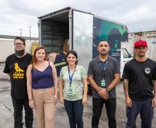 #praquemnaopodever cinco pessoas lado a lado posam para a foto em frente a um caminhão azul e verde que está com as portas da caçamba aberta e cheia de caixas com roupas.