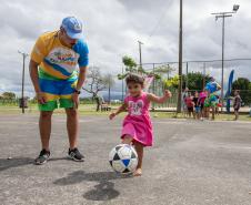 #praquemnaopodever uma criança de dois anos com um vestido rosa, chuta uma bola de futebol enquanto um representante do Governo do Paraná, com uma camisa colorida do verão maior, observa do lado esquerdo, encurvado, observando o chute da criança. ao fundo pessoas interagem no Aeroparque de Paranagua
