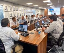 #ParaQuemNaoPodeVer A imagem mostra uma foto de uma reunião em uma sala de conferências. Há várias pessoas sentadas ao redor de uma mesa de madeira, todas olhando para a frente, onde parece haver uma apresentação ou videoconferência em andamento. Na parede ao fundo, há várias fotos emolduradas. As pessoas estão usando roupas formais, como camisas sociais. Na mesa, há laptops, garrafas de água e outros materiais de escritório. A sala é bem iluminada, com luzes no teto.