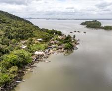 #ParaQuemNaoPodeVer A imagem mostra uma vista aérea de uma área costeira com vegetação densa e casas próximas à água. Há uma pequena península ou ilha coberta de árvores e algumas construções. A água ao redor é calma, e há algumas pequenas embarcações ou estruturas flutuantes próximas à costa. O céu está parcialmente nublado.