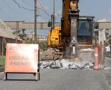 #ParaQuemNaoPodeVer A foto exibe uma obra na rua. Na imagem, há uma placa de aviso com o texto "ATENÇÃO OBRAS À FRENTE". Ao fundo, é possível ver uma escavadeira amarela e uma cerca de proteção laranja. No canto superior direito, há uma placa de trânsito indicando o limite de velocidade de 30 km/h. A cena parece estar em uma área urbana com calçadas e postes de eletricidade.