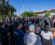 #ParaQuemNaoPodeVer A foto mostra um grupo grande de pessoas reunidas ao ar livre, em um local que parece ser uma área urbana com árvores e um prédio ao fundo. As pessoas estão em fila ou aglomeradas, e o céu está claro e azul.
