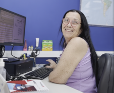 #praquemnaopodever Uma mulher de cabelos negros e lisos, regata lilas e óculos sorri para a camera. Ela está sentada em frente ao computador, mas precisou virar de lado para posar pra foto. O ambiente é de escritório.