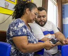 #praquemnaopodever uma mulher negra de blusa branca com plantas azuis mexe no celular ao lado de um rapaz de camisa branca com listras azul e laranja. Os dois estão sentados dentro de uma sala de aula e olham para o aparelho, enquanto a senhora de meia idade e óculos digita no celular.