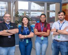 #ParaQuemNaoPodeVer Imagem exibe quatro pessoas posando juntas. As pessoas estão em pé, duas na frente e duas atrás, todas com os braços cruzados. A pessoa à esquerda, na parte de trás, é um homem usando uma camisa preta com detalhes em azul e jeans. Ele tem cabelo curto e barba. Ao lado dele, à direita, está uma mulher com cabelo escuro e liso, vestindo uma camisa azul. Na frente, à esquerda, está uma mulher com cabelo preso, usando uma blusa vermelha. À direita, na frente, está um homem com cabelo escuro 