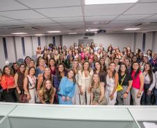 #ParaQuemNaoPodeVer Imagem mostra um grande grupo de pessoas, principalmente mulheres, posando juntas em um ambiente interno, possivelmente uma sala de conferências ou auditório. As pessoas estão sorrindo e parecem estar participando de um evento ou reunião. Algumas estão vestindo roupas coloridas e outras em tons mais neutros. 