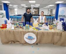 #ParaQuemNaoPodeVer A imagem mostra uma mesa coberta com uma toalha de tecido bege, decorada com coelhos de Páscoa e diversos produtos organizados em cima, como caixas de chocolate. Duas pessoas estão atrás da mesa, um homem e uma mulher, ambos sorrindo. Ao fundo, há um banner com o logotipo "Portos do Paraná" e uma parede azul. Há também um cartaz com uma foto e texto em preto e branco na parede. 