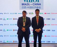 #ParaQuemNaoPodeVer A imagem mostra dois homens de terno em pé, lado a lado, em um evento. O fundo tem um banner com logotipos de várias empresas e as palavras "SUMMIT Valor Econômico BRAZIL-CHINA". Há também caracteres chineses no banner. Os homens estão de frente para a câmera, um deles segurando um microfone.