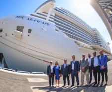 #ParaQuemNaoPodeVer Um grupo de oito pessoas está posando para uma foto em frente a um grande navio de cruzeiro. O navio tem o nome "MSC SEASCAPE" escrito na lateral. As pessoas estão vestidas formalmente, com ternos e camisas sociais. O ambiente parece ser um porto, com o navio atracado ao fundo.