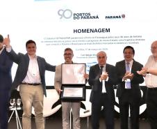 #praquemnaopodever homem te terno cinza e óculos de grau segura uma caixa aberta com uma placa dentro.  Ele e outros cinco homens brancos de roupa social estão em cima de um palco, alguns sorriem, outroas fazem joinha. Ao fundo, aparece escrito "Homenagem 90 anos" no telão