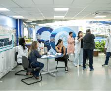 #ParaQuemNaoPodeVer Um grupo de pessoas está em um ambiente interno que parece ser uma feira ou evento de negócios. Há uma mesa com duas mulheres sentadas, uma delas usando jaleco branco e a outra com cabelo comprido e loiro. Elas estão interagindo com outras pessoas em pé. No fundo, há um banner com o texto "Portos do Paraná" e imagens de paisagens. Algumas pessoas estão vestindo jalecos brancos, sugerindo que podem ser profissionais de saúde ou de outra área técnica. O ambiente é bem iluminado, com teto d