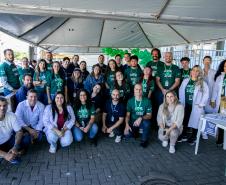 #ParaQuemNaoPodever Um grupo grande de pessoas está posando para uma foto em um ambiente externo. A maioria delas está usando camisetas verdes com a inscrição "ABRIL VERDE 2025". Algumas pessoas estão agachadas na frente, enquanto outras estão em pé atrás. Há uma tenda ao lado direito da imagem e algumas bexigas verdes. No canto inferior esquerdo, há uma mesa branca com alguns objetos em cima, incluindo um livro amarelo e um panfleto azul.