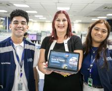 #ParaQuemNaoPodeVer Três pessoas estão sorrindo para a câmera em um ambiente interno, possivelmente um escritório. A pessoa à esquerda é um jovem de cabelo curto e escuro, vestindo um colete azul com a palavra "ESTAGIÁRIO" escrita. Ele também usa um crachá e um cordão azul. No centro, uma mulher de cabelo ruivo segura um certificado ou placa azul, e está vestindo uma blusa preta. À direita, uma mulher de cabelo castanho comprido veste um colete azul semelhante ao do jovem, com a inscrição "Portos do Paraná"