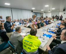 #praquemnaopodever setenta pessoas estão sentadas olhando para um senhor em pé, de camisa azul escura e calça bege. Ele segura um microfone na mão e com olhar focado por trás dos óculos, observa o público que o acompanha. A sala está com as cortinas brancas fechadas e ao fundo a imprensa grava o encontro.