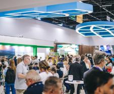 #ParaQuemNaoPodeVer Foto exibe um evento ou feira com muitas pessoas. Há uma estrutura grande e moderna no teto, com formas geométricas azuis. As pessoas estão em pé e algumas estão sentadas em mesas, parecendo interagir ou observar algo. O ambiente está movimentado e há uma variedade de pessoas, algumas com mochilas. 