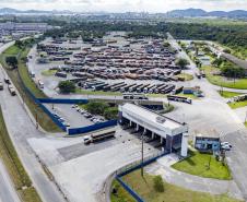 #ParaQuemNaoPodeVer Imagem aérea de um grande pátio industrial ou logístico. No centro da imagem há um prédio principal de formato retangular, com várias vagas de estacionamento ao redor. À direita do prédio, há uma grande quantidade de caminhões ou carretas estacionados em fileiras organizadas. O local é cercado por áreas verdes e ruas asfaltadas, com alguns veículos circulando. Ao fundo, é possível ver uma cidade e montanhas.