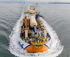 #ParaQuemNaoPodeVer Imagem aérea de um navio em movimento no mar. O navio está cortando a água, criando uma esteira branca ao seu redor. A embarcação possui várias estruturas e equipamentos coloridos em sua parte superior, incluindo elementos em azul, laranja e amarelo.