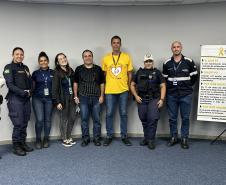 #ParaQuemNaoPodeVer Foto de um grupo de oito pessoas, sendo quatro delas usando uniformes de agentes de trânsito ou policiais, e as outras quatro usando roupas casuais. Eles estão em pé, lado a lado, em frente a uma parede clara e um tapete azul. À frente do grupo, há um banner com informações sobre o movimento Maio Amarelo, que é uma campanha de conscientização para a segurança no trânsito. O banner tem detalhes em amarelo e branco, com o símbolo do laço amarelo. Todos estão sorrindo e olhando para a câmer