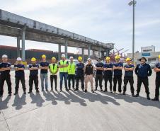 #ParaQuemNaoPodeVer Foto de um grupo de pessoas em um porto. Elas estão alinhadas, usando capacetes de segurança amarelos e brancos, e algumas vestem coletes refletivos. Ao fundo, há uma estrutura de concreto que de carregamento de granéis.