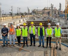 #ParaQuemNaoPodeVer Grupo de oito pessoas em um canteiro de obras. Todos estão usando equipamentos de segurança, como capacetes e coletes refletivos. A maioria veste coletes amarelos, enquanto uma pessoa usa um colete laranja. O cenário ao fundo mostra estruturas de concreto e uma grua amarela, indicando que estão em uma área de construção. O céu está nublado e há postes de eletricidade ao fundo.