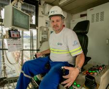 #ParaQuemNaoPodeVer Foto de um homem sentado em uma cabine de operação. Ele está usando capacete branco com o logotipo da empresa "ICTSI" e uniforme azul com faixas refletivas amarelas. O homem está sentado em uma cadeira diante de um painel de controle com vários botões coloridos e alavancas. À frente dele, há uma tela de computador com gráficos e informações técnicas. A cabine tem janelas grandes, permitindo ver estruturas metálicas e o que parece ser um terminal portuário ao fundo. O ambiente sugere que 