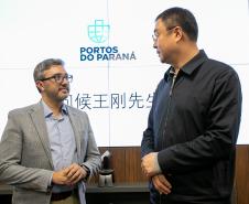 #praquemnaopodever na foto, dois homens conversam em frente a uma tela branca com logo escrita "Portos do Paraná" e dizeres em mandarim embaixo. A esquerda, o homem branco de terno cinza, camisa azul e óculos segura as mãos enquanto conversa com um homem chinês mais alto, de óculos, camisa e calça pretas.