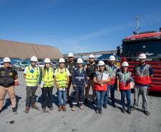 #ParaQuemNaoPodeVer Imagem mostra grupo de pessoas posando em frente a um caminhão vermelho. Todos estão usando capacetes de segurança e coletes refletivos, indicando que podem estar em um ambiente de trabalho ou construção. Ao fundo, há um grande galpão e o céu está azul.