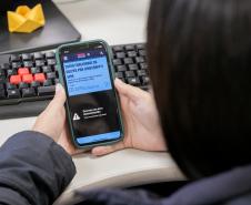 #ParaQuemNaoPodeVer Imagem mostra uma foto de uma pessoa segurando um celular. O celular está exibindo uma página da internet com o título “COMO FUNCIONAM OS GOLPES POR WHATSAPP E SMS”. Abaixo do título, há um texto menor, mas não está totalmente legível na imagem. O celular está em cima de um teclado de computador com algumas teclas vermelhas e outras pretas. No canto superior direito da mesa, há um objeto amarelo que parece ser uma dobradura de papel (origami). A pessoa está usando uma blus