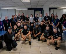 #ParaQuemNaoPodeVer Foto de grupo em ambiente de escritório. Aproximadamente vinte pessoas estão reunidas, a maioria usando camisetas pretas com logotipos e calças bege, sugerindo que fazem parte de uma equipe ou evento. Algumas pessoas estão de crachá e há diversidade de gênero e idade. Todos estão sorrindo e posando para a foto, alguns agachados na frente e outros em pé atrás. Ao fundo, há divisórias de escritório, mesas e cadeiras.