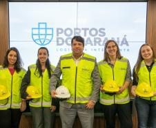 #ParaQuemNaoPodeVer Foto de cinco pessoas em pé, todas usando coletes de segurança amarelo e segurando capacetes (a maioria amarelos, um branco). Elas estão sorrindo e posando para a foto em frente a um painel com o logotipo azul e as palavras “PORTOS DO PARANÁ LOGÍSTICA INTELIGENTE”. O grupo é composto por quatro mulheres e um homem, todos com aparência profissional.