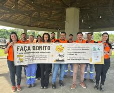 #ParaQuemNaoPodeVer Grupo de pessoas vestindo camisetas laranja posando para uma foto. Eles estão segurando um cartaz que diz: "FAÇA BONITO. Proteja nossas crianças e adolescentes. 25 anos. Dia Nacional de Combate ao Abuso e à Exploração Sexual de Crianças e Adolescentes". O cartaz também menciona o Disque 100 e fala sobre a importância de proteger crianças e adolescentes. Ao fundo, há uma estrutura coberta e uma área verde.