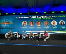 #ParaQuemNaoPodeVer Palco de conferência com seis pessoas sentadas em cadeiras, participando de um painel de discussão. Ao fundo, há um grande painel azul com a imagem de um contêiner portuário e o título do evento em espanhol: "Expansión portuaria en América Latina: casos de éxito y retos estratégicos". Abaixo do título, aparecem fotos e nomes dos participantes do painel, com seus respectivos cargos e empresas. O ambiente é formal, com todos vestidos de maneira profissional. O palco está bem iluminado e há