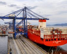 #ParaQuemNaoPodeVer Imagem de um navio cargueiro vermelho atracado em um porto. O navio está carregado com vários contêineres empilhados e há guindastes azuis grandes posicionados para carregar ou descarregar os contêineres. À esquerda, é possível ver parte do cais com caminhões e veículos pequenos. O mar está calmo e o céu está parcialmente nublado.