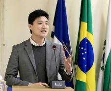 #ParaQuemNaoPodeVer foto de um homem jovem, de pele clara e cabelo preto, vestindo terno cinza, suéter escuro e camisa branca. Ele está em pé atrás de um púlpito, falando ao microfone, com expressão sorridente. Atrás dele, há bandeiras, incluindo a do Brasil e outra azul com a palavra "NITOINI" parcialmente visível. O ambiente parece ser formal, possivelmente uma apresentação ou discurso.