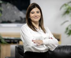 #praquemnaopodever foto de uma mulher de camisa branca, braços cruzados e semblante sorridente. Ao fundo, uma parede branca com o mapa aéreo dos portos paranaense, plantas, um sofá preto e uma mesa marrom vista de lado, mas que sustenta um mapa interativo dos portos paranaenses.