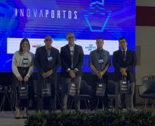 #ParaQuemNaoPodeVer Na imagem do evento, há cinco pessoas (quatro homens e uma mulher) em pé, posando para foto em um palco. Todos estão vestidos de forma formal, com crachás pendurados no pescoço. Eles seguram sacolas pretas com o logo do evento. Ao fundo, há um painel azul grande com a palavra "INOVAPORTOS" escrita na vertical. Também é possível ver algumas cadeiras vazias no palco e plantas decorativas na frente. O ambiente parece ser de uma conferência ou seminário.