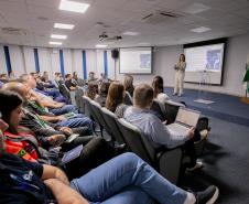 #ParaQuemNaoPodeVer Auditório com várias pessoas sentadas em cadeiras, assistindo a uma apresentação. No fundo da sala, uma mulher está em pé, falando ao microfone, com um telão atrás dela exibindo um slide. Há bandeiras do Brasil e de outros estados ou instituições ao lado do palco. As pessoas na plateia parecem estar prestando atenção, algumas com crachás e blocos de anotações. O ambiente é iluminado por luzes de teto e tem paredes brancas com detalhes em azul.