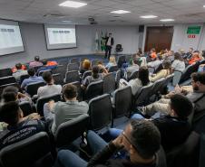 #ParaQuemNaoPodeVer Sala de aula ou auditório com várias pessoas sentadas em cadeiras, todas voltadas para a frente, assistindo a uma apresentação. Na frente da sala, há uma pessoa de pé, aparentemente palestrando, e dois telões com slides projetados na parede. No canto direito, há três bandeiras, incluindo a do Brasil. Muitas pessoas estão usando camisetas cinza com a inscrição “AUTORIDADE PORTUÁRIA PORT AUTHORITY” nas costas. O ambiente parece formal e voltado para treinamento ou reunião.
