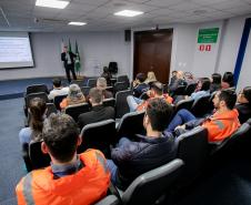 #ParaQuemNaoPodeVer Sala de reuniões vista de trás, com várias pessoas sentadas em cadeiras, assistindo a uma apresentação. Muitas dessas pessoas estão usando uniformes laranja com faixas refletivas, típicos de trabalhadores de áreas industriais ou de segurança. Na frente da sala, há uma pessoa em pé, aparentemente apresentando um slide projetado na parede. Ao lado do apresentador, estão duas bandeiras (provavelmente do Brasil e do estado). A sala tem piso azul, paredes claras e teto com iluminação embutida