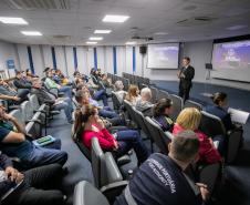 #ParaQuemNaoPodeVer Auditório com várias pessoas sentadas em cadeiras, todas de cabeça baixa. Na frente, um homem de terno está em pé, também de cabeça baixa, próximo a um púlpito transparente. Ao fundo, há dois telões projetando a mesma imagem, com fundo roxo e texto branco, mas o texto não está totalmente legível na foto. O ambiente é fechado, com iluminação de teto e paredes claras.