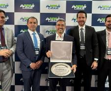 #ParaQuemNaoPodeVer Foto de cinco homens de terno, posando para uma foto em frente a um painel com o logo “ANDAV LATAM”. Eles estão sorrindo e segurando um quadro e um troféu, sugerindo que receberam algum tipo de prêmio ou reconhecimento. O ambiente parece ser um evento corporativo ou uma premiação.
