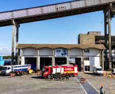 #ParaQuemNaoPodeVer  foto de um pátio industrial ou de uma garagem de veículos grandes. No centro da imagem, há dois caminhões: um caminhão de bombeiros vermelho com a inscrição "BOMBEIROS" e outro caminhão branco com detalhes azuis, que parece ser de resgate ou suporte. Ambos estão estacionados próximos a um prédio de estrutura robusta, com cobertura alta e colunas de concreto. Há cones e barreiras laranja ao redor dos veículos, indicando uma área isolada. Algumas pessoas com coletes refletores estão próxi