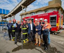 #praquemnaopodever Na foto um grupo de homens faz o sinal de jóia com o polegar e sorriem em frente a um caminhão de bombeiros. Ao fundo, um predio branco e dalas suspensas que conectam empresas ao cais.