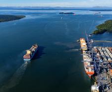 #ParaQuemNaoPodeVer Imagem aérea de um porto mostrando um grande navio saindo do cais, cercado de contêineres e equipamentos portuários. O mar, de azul intenso, ocupa a maior parte da imagem, com algumas pequenas ilhas verdes ao fundo. Outros navios menores podem ser vistos mais afastados, navegando ao longe.